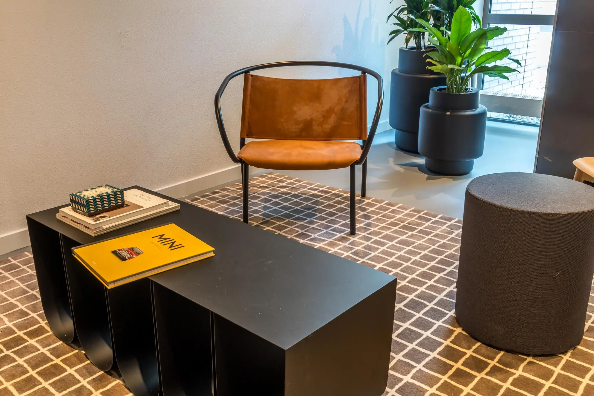 Moderne wachtruimte met een leren designstoel, zwarte salontafel met boeken en tijdschriften, grijs krukje en grote planten in zwarte potten.